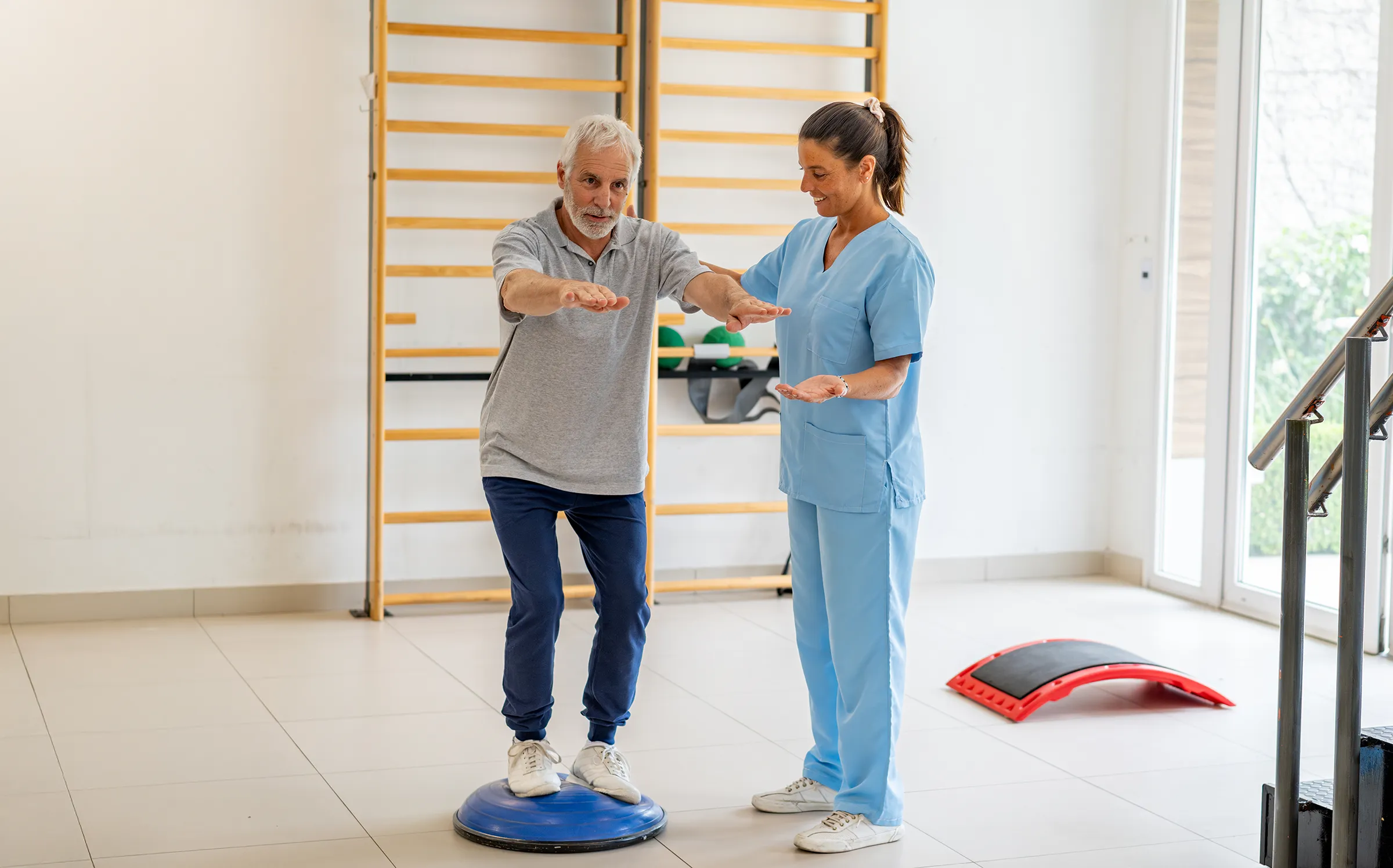 Healthcare professional assisting an older man with balance training on a stability trainer in a bright therapy room at The Birches of Leigh Valley.